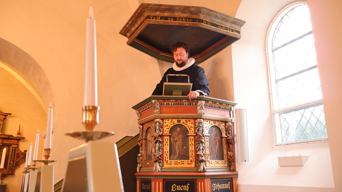 En person står ved en prægtig prædikestol i en kirke og læser op fra en bog under en gudstjeneste.