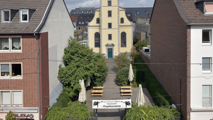 Eine charmante Stadtansicht mit einem hohen Uhrturm, gesäumt von malerischen Gebäuden und einem Schild für ein Freiluft-Orgelkonzert.