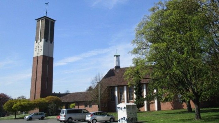 Eine friedvolle Kirche mit einem hohen Uhrturm und einer Backsteinfassade, umgeben von geparkten Autos und üppigem Grün.