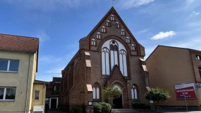 Eine Backsteinkirche mit hohen Spitzbogenfenstern und einem steilen Satteldach steht markant an einer Straßenecke.