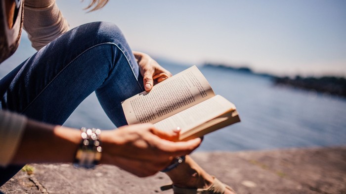 Eine Person sitzt am Wasser und liest ein Buch.