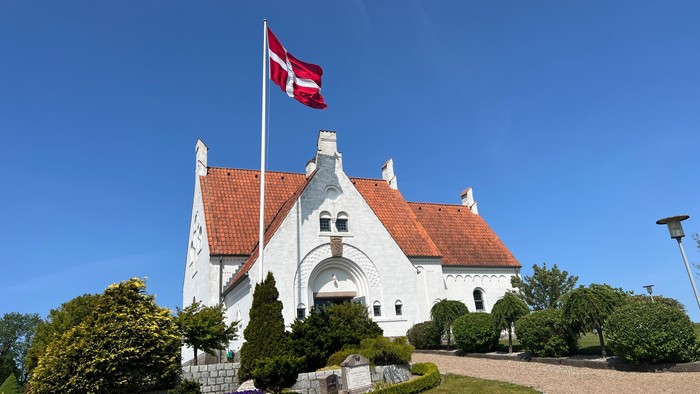 Et malerisk hvidt hus med rødt teglstenstag, hvor et dansk flag vajer under en klar blå himmel.