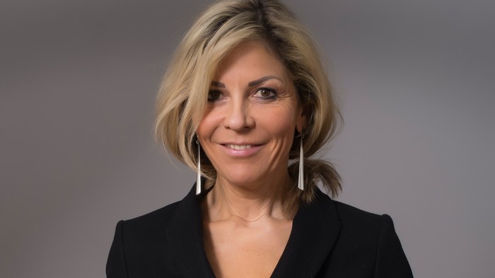 Professionelle Frau in einem schwarzen Blazer, die mit neutralem Hintergrund in die Kamera lächelt.
