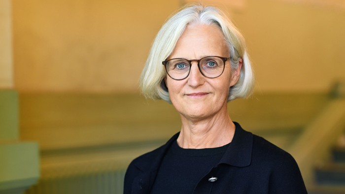 Eine lächelnde Frau mit kurzem grauem Haar und Brille steht vor einer strukturierten Wand.
