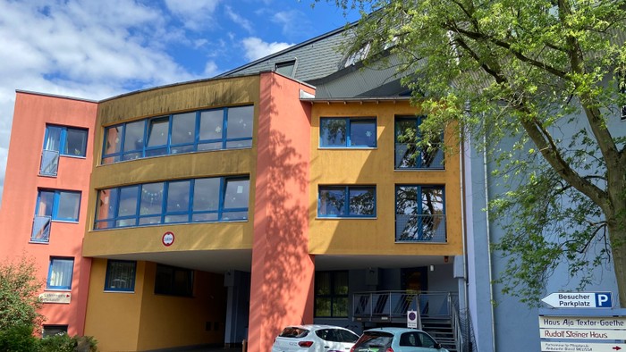 Bild des Aja Textor Haus in Frankfurt Eschersheim von der Einfahrt her gesehen. Das Gebäude ist oder, rose und hellblau gestrichen.