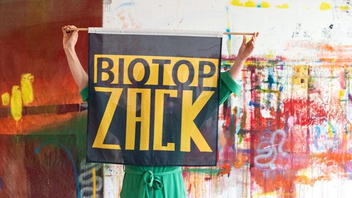 Person in grüner Hose hält ein Schild mit der Aufschrift „BIOTOP ZHLK“ in einem mit Kunst gefüllten Atelierraum.