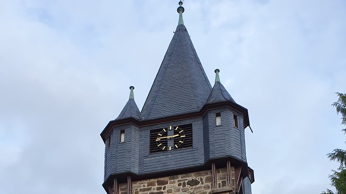Historischer Steinturmuhr mit spitzem Dach und aufwendigen Mauerwerksdetails in einer parkähnlichen Umgebung.