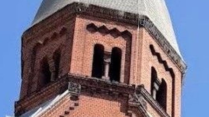 Historische Backsteinkirche mit hohem Turm und gewölbtem Eingang, umgeben von Bäumen unter klarem Himmel.