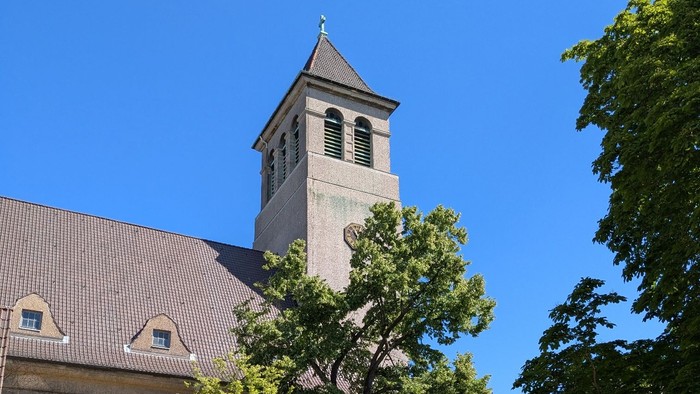 Kirchturm mit Uhr und Kreuz, umgeben von Bäumen und Gebäuden