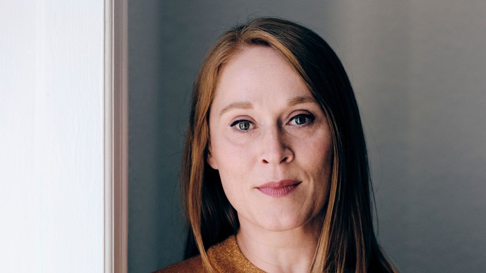 En kvinde i en sennepsfarvet sweater står selvsikkert ved et vindue med en neutral baggrund.