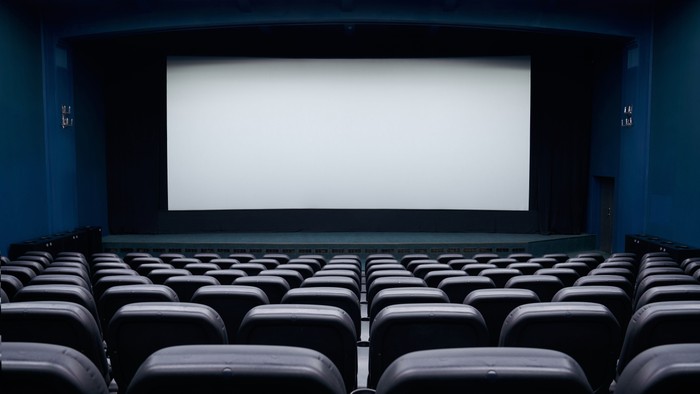 Ein leeres Kino mit vielen schwarzen Sitzen und einer großen weißen Leinwand.