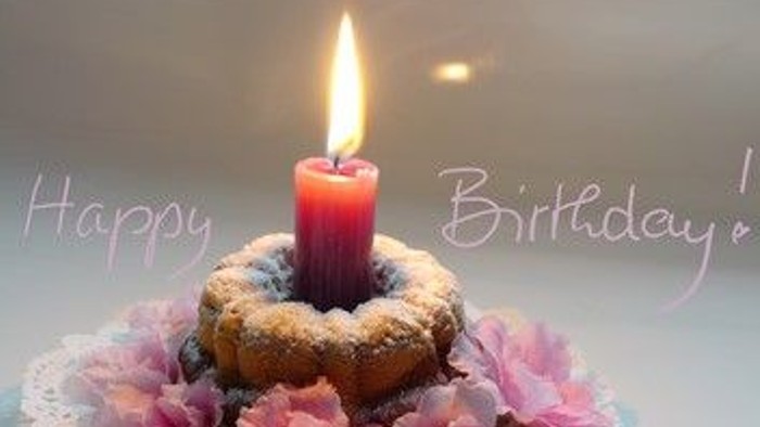 Ein festlicher Geburtstags-Donut mit einer brennenden Kerze und rosa Blumen auf einer Spitzenunterlage, mit den Worten „Alles Gute zum Geburtstag!“.