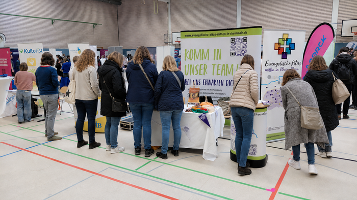 Zahlreiche Menschen informieren sich am Messestand der Evangelischen Kitas über berufliche Möglichkeiten. Roll-ups und Informationsmaterialien werben unter dem Motto „Komm in unser Team!“ für die Arbeit in den Kitas.