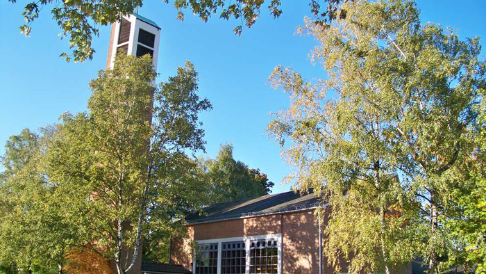 Eine malerische Backsteinkirche mit einem hohen Glockenturm, umgeben von üppigen Herbstbäumen unter einem klaren blauen Himmel.