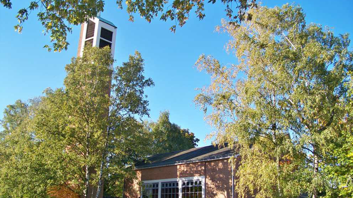 Eine malerische Backsteinkirche mit einem hohen Glockenturm, umgeben von üppigen Herbstbäumen unter einem klaren blauen Himmel.