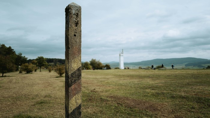 Verwitterter Pfosten steht markant auf einem offenen Grasfeld mit fernen Hügeln und einem einsamen Denkmal.