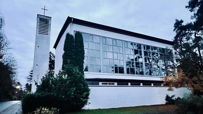 Moderne Kirchengebäude mit großen Glasfenstern und einem hohen Glockenturm neben üppigem Grün.