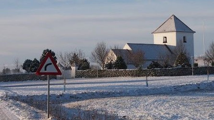 Sneklædt mark med trekantet advarselsskilt, stendige og kirke i baggrunden