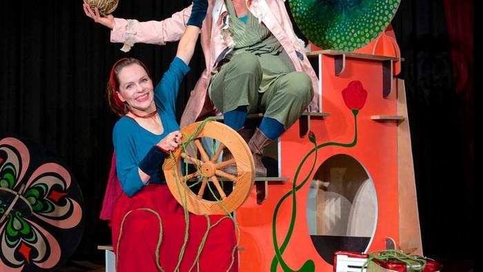 Verspieltes Puppentheater mit Schauspielern, übergroßen Requisiten und einem Fantasieschloss-Bühnenbild.