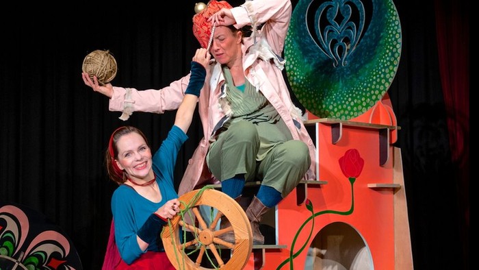 Verspieltes Puppentheater mit Schauspielern, übergroßen Requisiten und einem Fantasieschloss-Bühnenbild.