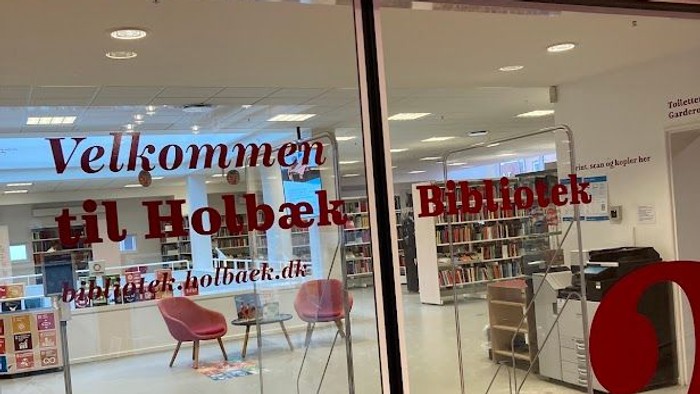 Velkomstskilt til Holbæk Bibliotek med moderne indretning og siddeområde synligt gennem glasdørene.