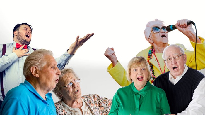 Eine Gruppe älterer Menschen feiert ausgelassen, einer hält ein Mikrofon, eine Frau liest aus einem Buch vor.