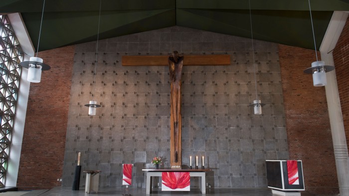 Die Kirche zeigt einen Altar mit Kreuz und Kerzen, flankiert von zwei roten Teppichen und einem Bildschirm.