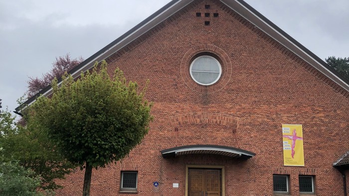 Ziegelsteinkirche mit zentralem Eingang, kleinem Turm und einem bunten Banner über der Tür.