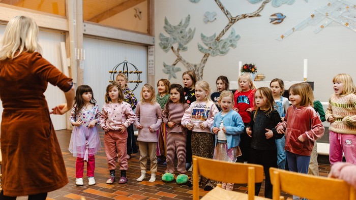 Børn synger sammen i en kirke, mens en voksen leder, omgivet af dekorativ vægkunst.