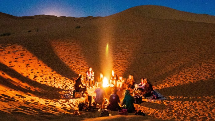 Eine Gruppe von Menschen sitzt um ein Lagerfeuer in einer Wüste unter dem Sternenhimmel.