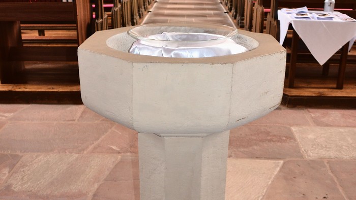 Steinerner Taufstein in einem Kirchenschiff, das zum Altar führt, mit hölzernen Kirchenbänken.