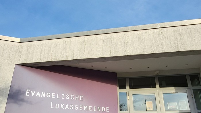 Moderner Kircheneingang mit der Beschilderung „Evangelische Lukasgemeinde“, klarer Himmel und Topfpflanzen in der Nähe.