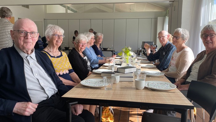 En gruppe ældre mennesker samlet omkring et langt bord i et lyst indendørs spiseområde.