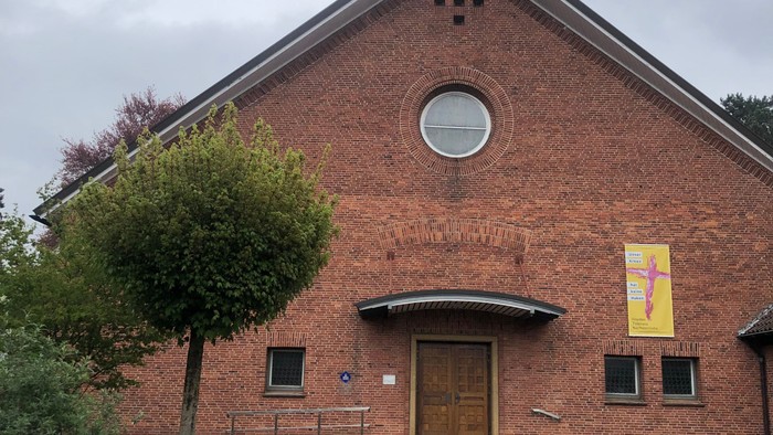 Ziegelsteinkirche mit zentralem Eingang, kleinem Turm und einem bunten Banner über der Tür.