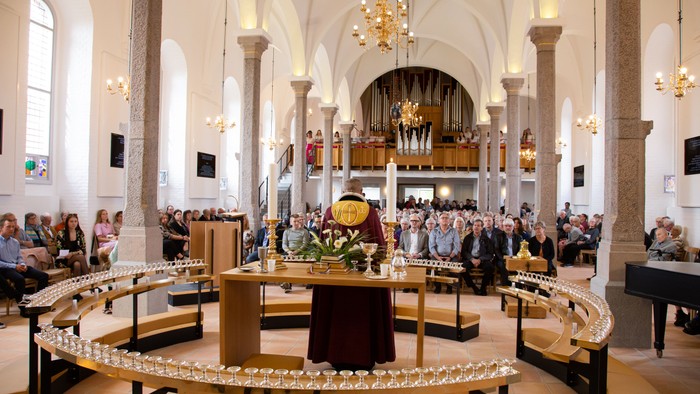 Inden i en stor kirke med høje buer og lyse vægge, sidder en gruppe af mennesker i en halocirkel om et bord med blomster og lys.