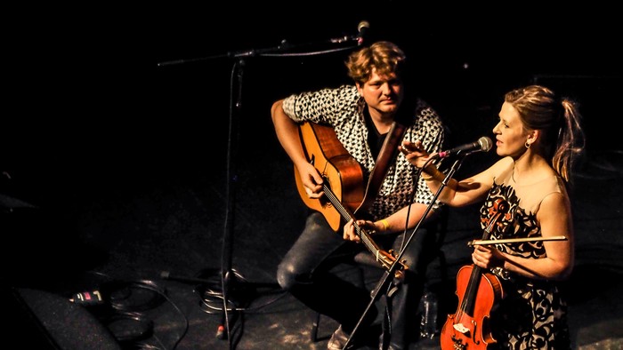To musikere optræder live på scenen, den ene spiller guitar og den anden violin med en mikrofon.