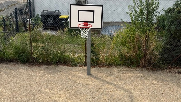 Auf dem Foto sieht man mit Blick von schräg oben nach unten einen Bolzplatz mit Basketballkorb in der Mitte. Im Hintergrund steht ein weiß gestrichenes Haus.