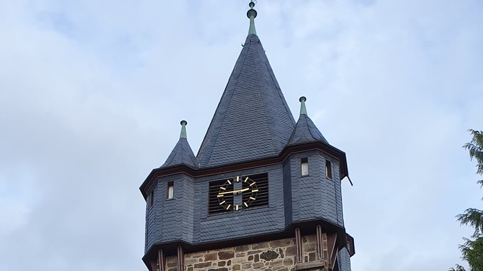 Historischer Steinturmuhr mit spitzem Dach und aufwendigen Mauerwerksdetails in einer parkähnlichen Umgebung.