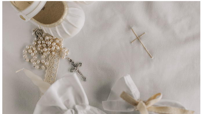 A white cloth embroidered with a cross; on top the cloth is a child's white satin shoe, the sleeve of a garment, a white tied pouch and a white rosery.