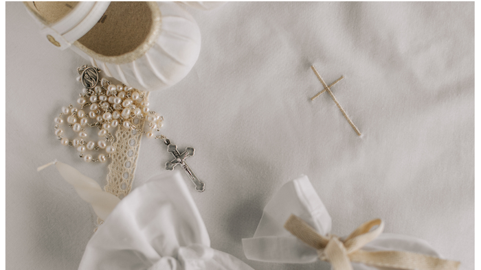 A white cloth embroidered with a cross; on top the cloth is a child's white satin shoe, the sleeve of a garment, a white tied pouch and a white rosery.