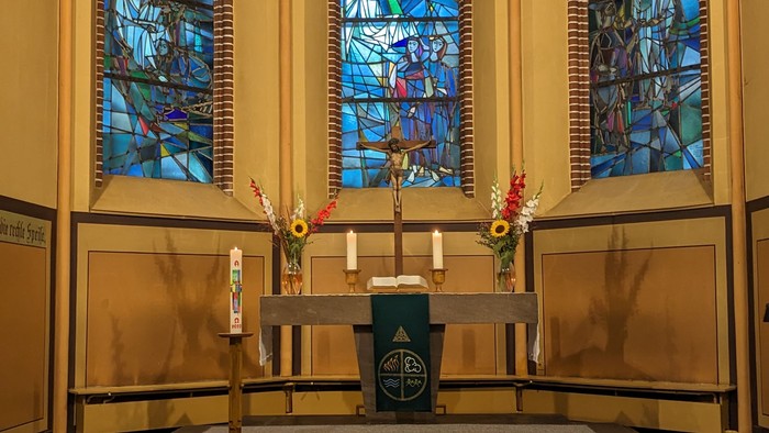 Apsis der Magdalenenkirche mit drei gestaltenten Fenstern und Altar mit Kreuz, Kerzen, Blumenschmuck und grünem Parament.