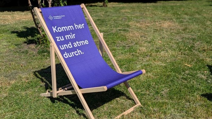Blauer Liegestuhl auf dem Rasen mit deutschem Text, der Menschen einlädt, zu kommen, zu entspannen und durchzuatmen.
