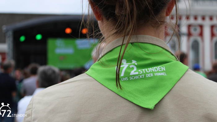 Person mit einem grünen „72 Stunden“-Schal bei einer Outdoor-Veranstaltung mit Menschenmenge und Bühne im Hintergrund.