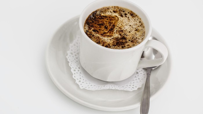 En skummende cappuccino i en hvid kop med en ske på et blonderunderlag og en underkop.
