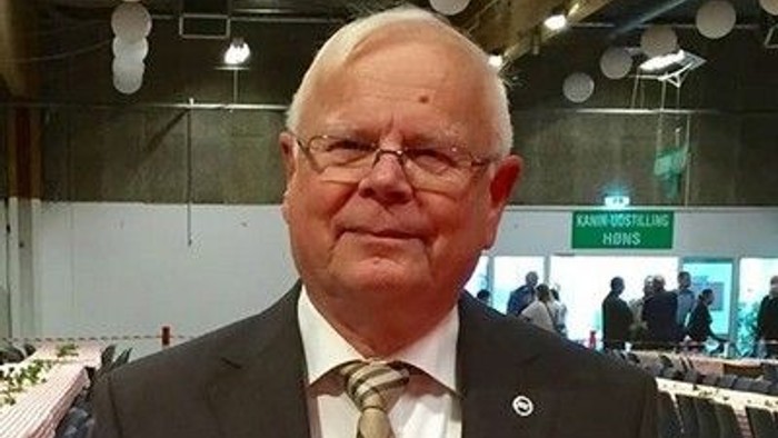En mand i formelt tøj med slips står til en indendørs begivenhed eller sammenkomst.