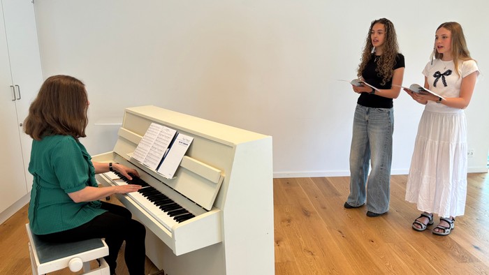En kvinde spiller piano mens to andre kvinder står ved siden af og læser fra noter.