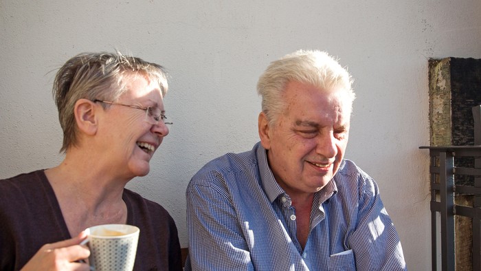 Zwei ältere Menschen lächeln, während sie gemeinsam drinnen Kaffee genießen.
