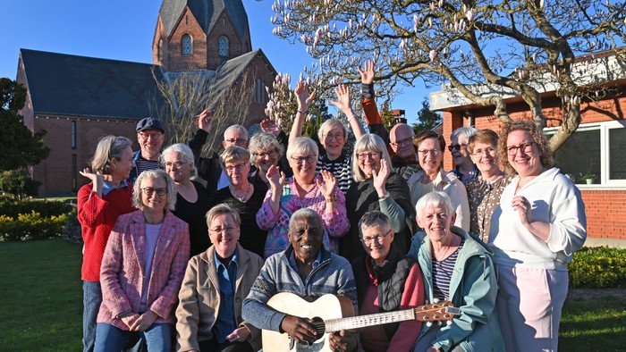 En glad gruppe poser udendørs nær en kirke, hvor én spiller guitar, og alle smiler bredt.
