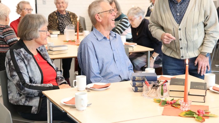 En gruppe ældre personer sidder ved bord i en lokal, hvor de ser ud til at have en fælles aktivitet.