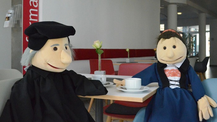 Zwei Puppen in historischer Kleidung posieren gemeinsam an einem kleinen Cafétisch mit Kaffee.